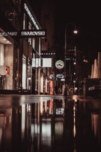 Street view of a lit shopping area in Vienna with reflections on wet pavement at night.